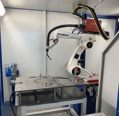 Braccio robotico industriale in funzione all'interno di un piccolo laboratorio, accanto a un tavolo da lavoro metallico.