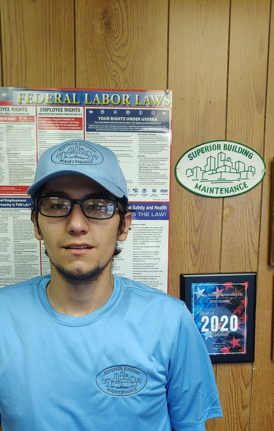 Person in glasses and blue shirt standing before a wood-paneled wall with signs and a 2020 poster.