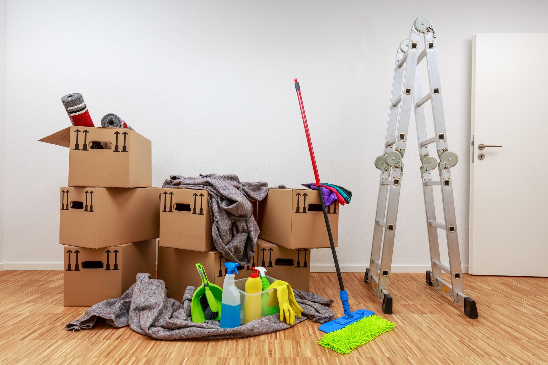 Cleaning supplies, mop, ladder, and moving boxes in a room with a wooden floor and white door.