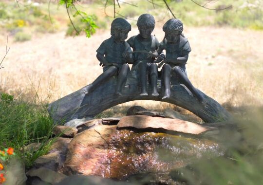 a statue of three children sitting on a bridge over a stream