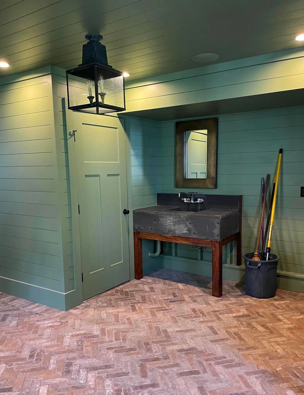 Green-painted room with stone sink on wooden legs, door, brick floor, lantern light, and cleaning supplies.