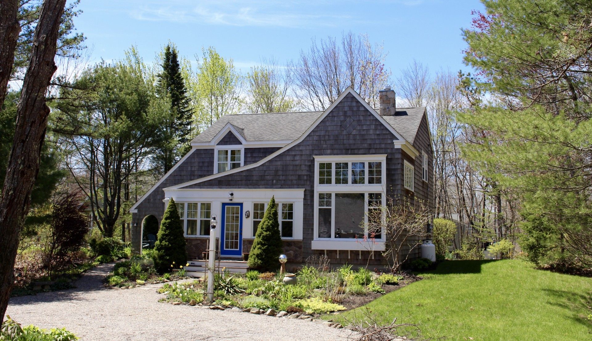 Gray-shingled house with blue door, white trim, and garden, surrounded by trees and a gravel driveway on a sunny day.