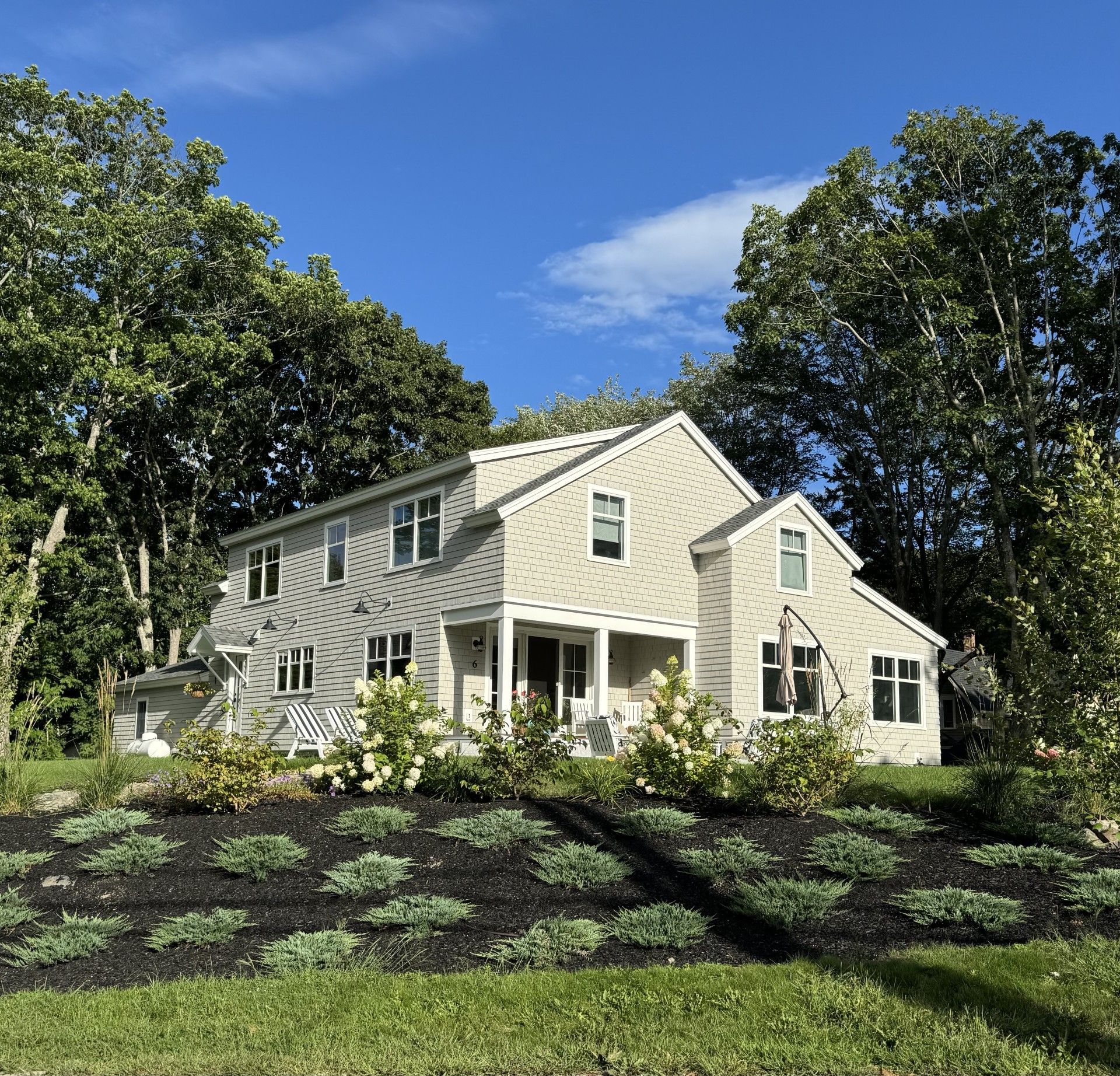 Two-story beige house with a porch and green landscaping under a blue sky.
