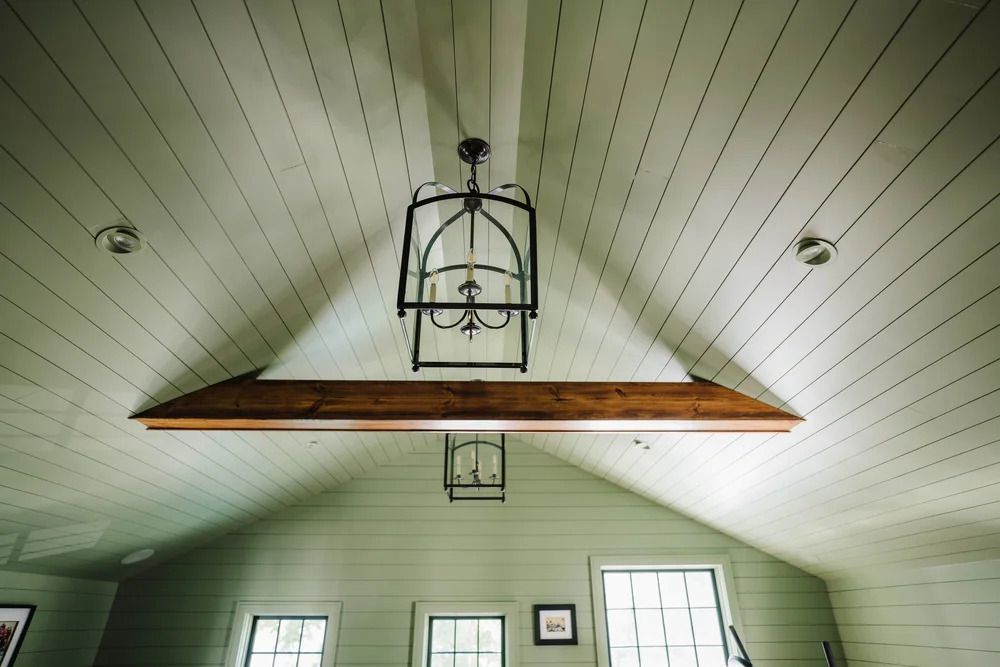 Light green sloped ceiling with dark wood beam and ornate chandelier. Small windows in room below.