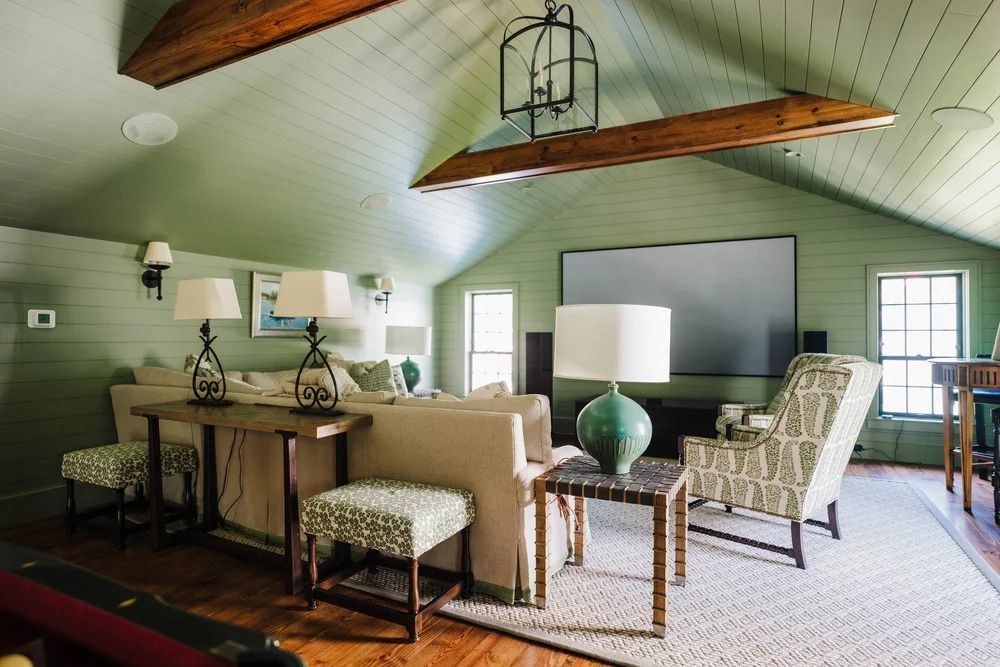 Green-walled living room with light-colored sofa, patterned chair, and a large screen. Wooden beams on ceiling.