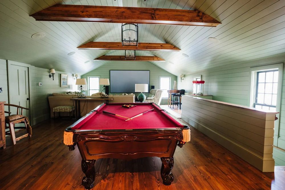 Pool table in a room with a sloped ceiling and exposed beams, a seating area visible in the background.