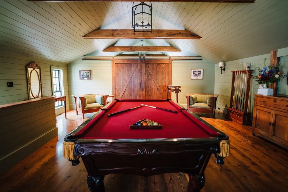 Pool table in a wood-paneled room. Red felt table, wooden beams, two chairs, and a bar-style light fixture.