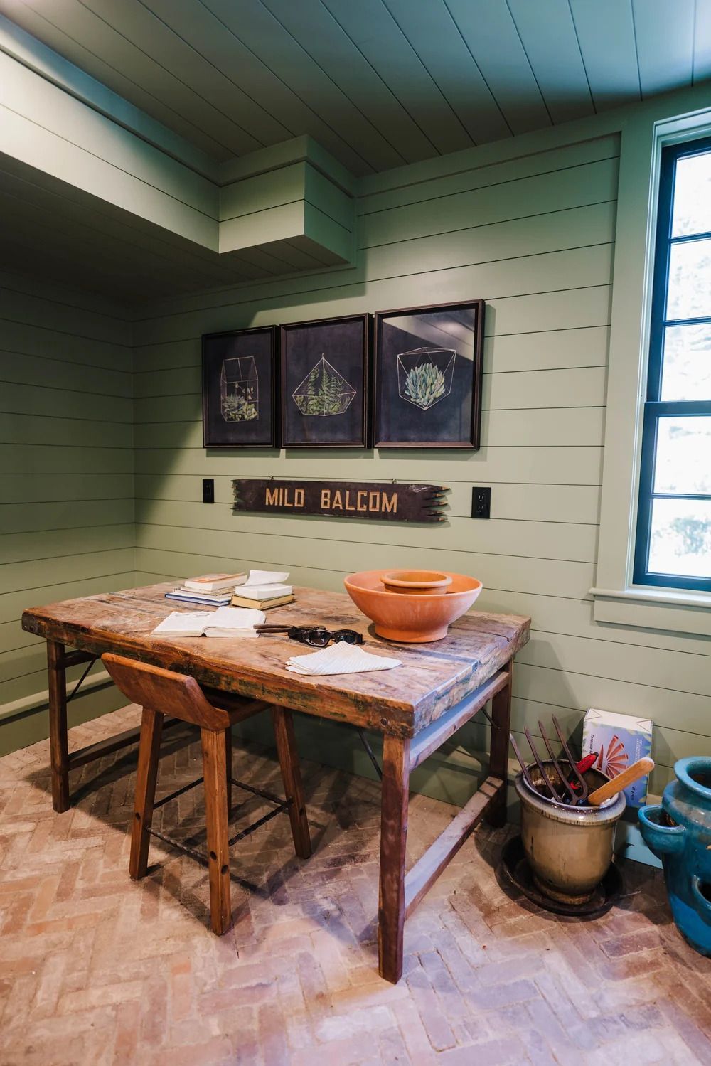 A rustic workspace with a wooden table, stool, and artwork against a green-painted paneled wall.