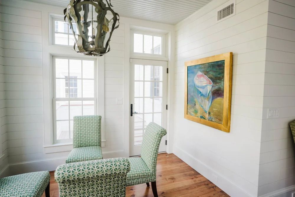 Bright room with white paneled walls, wood floor, and green patterned chairs; framed painting hangs on wall.