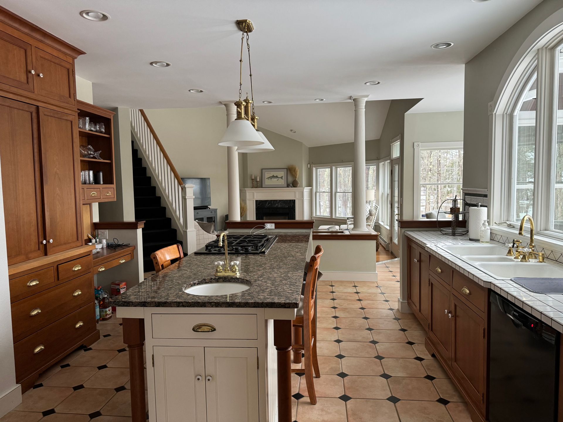 Spacious kitchen with wood cabinets, island, and tile floor, leading to a living area with fireplace and staircase.