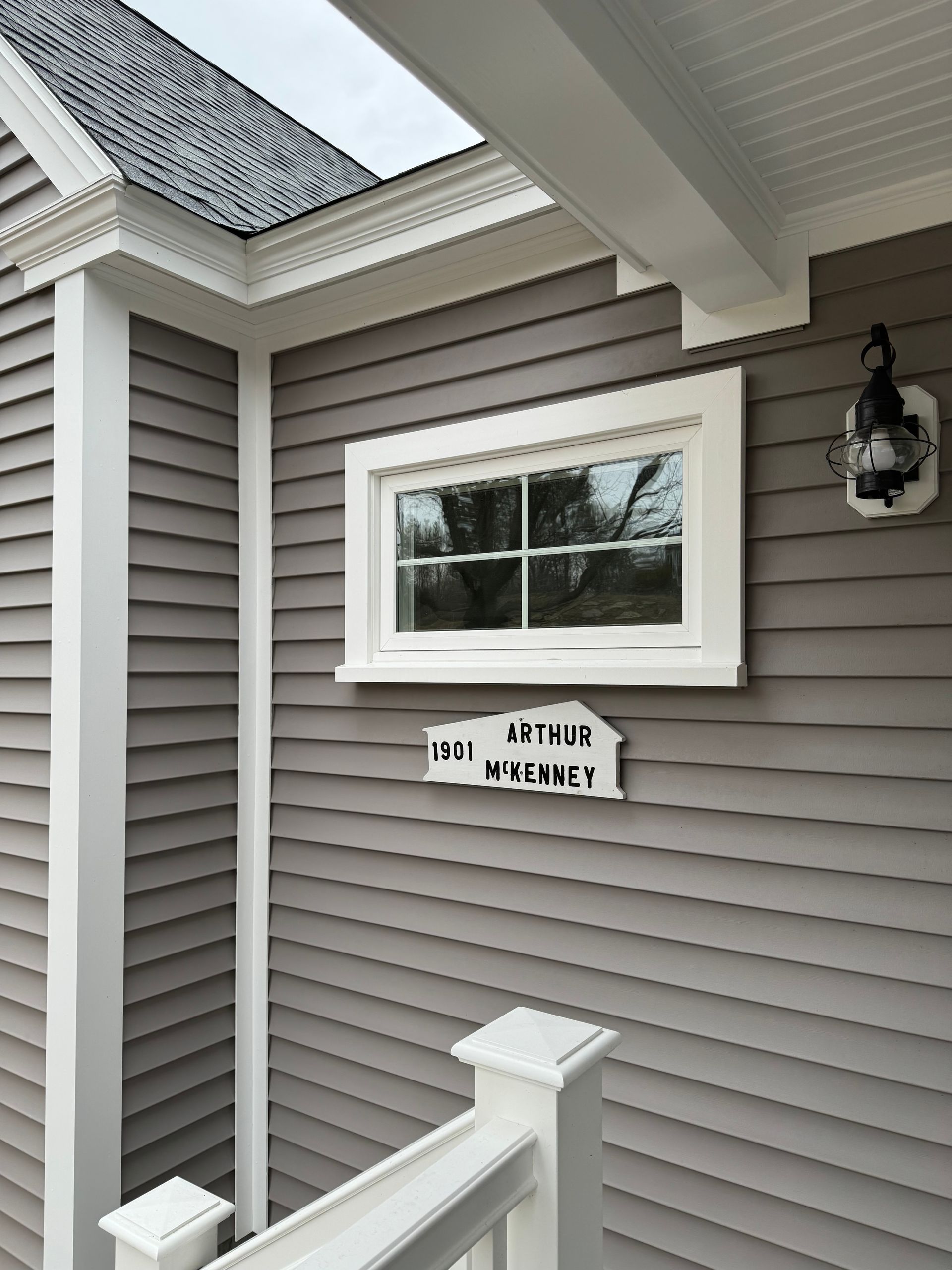 Gray house siding with a white-framed window. A house number plaque is below the window.
