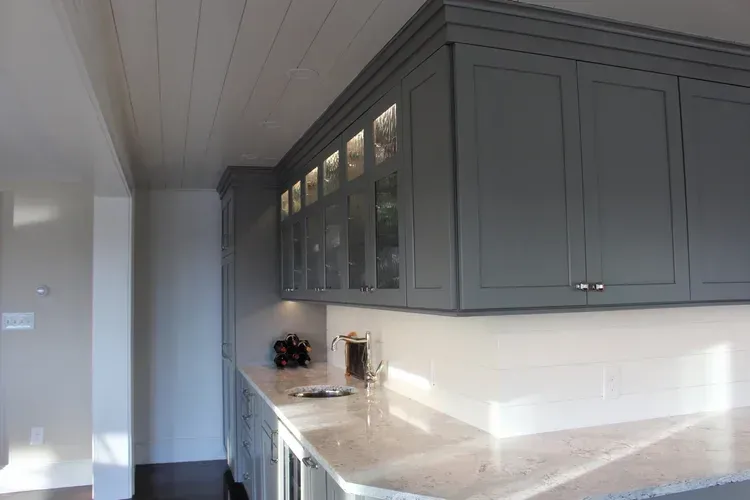 Gray kitchen cabinets with glass doors over a white countertop, wood ceiling.