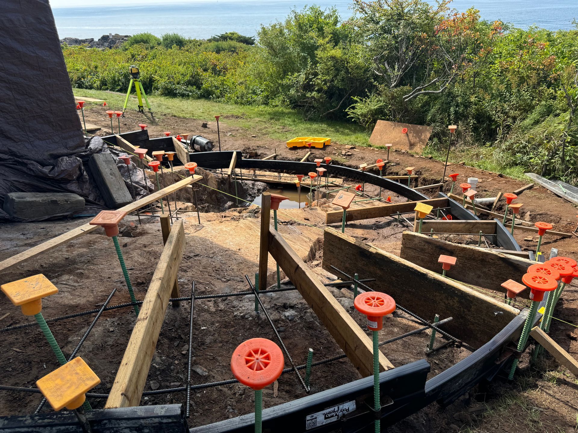 Construction site outdoors, concrete forms with rebar, orange stakes, and ocean view in background.