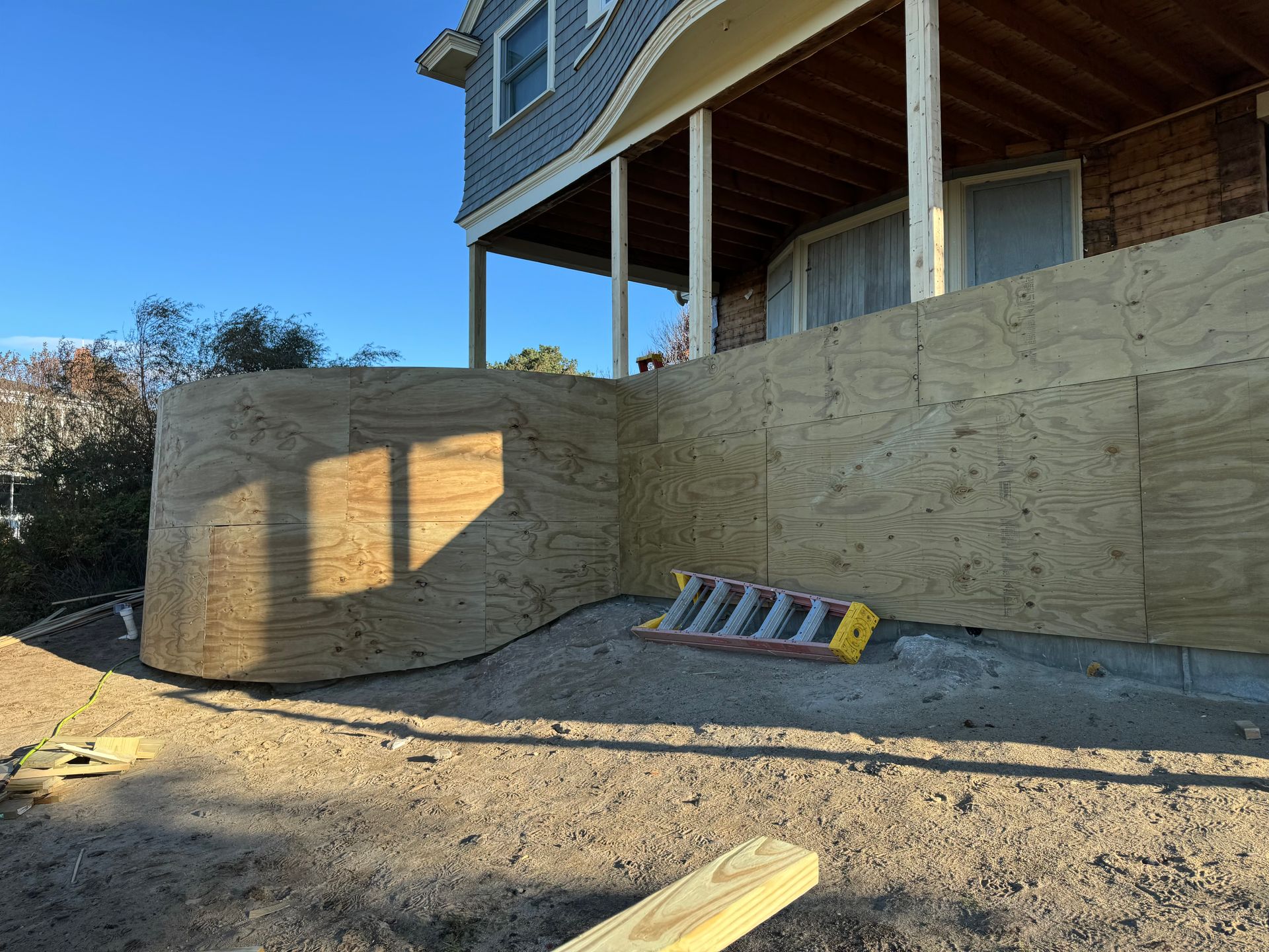 Exterior of a house under construction; plywood covers the base. A small ladder is present.