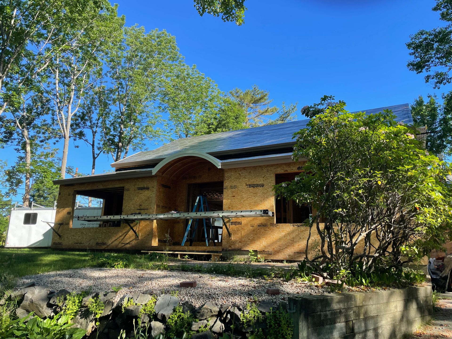 Unfinished wooden house under construction; trees in background, clear blue sky.
