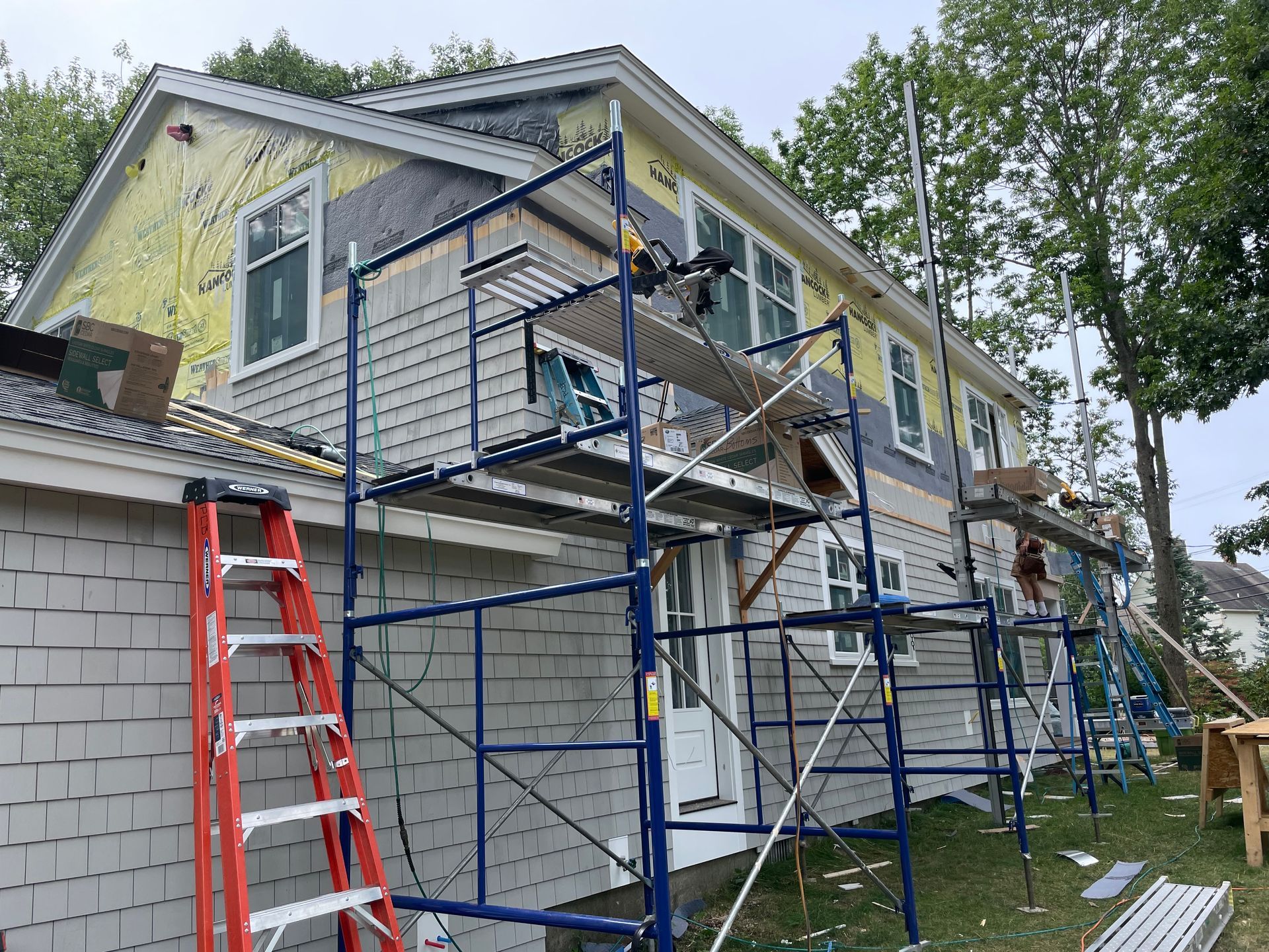 House under construction; siding installation with scaffolding, ladder, and partially installed gray shingles.