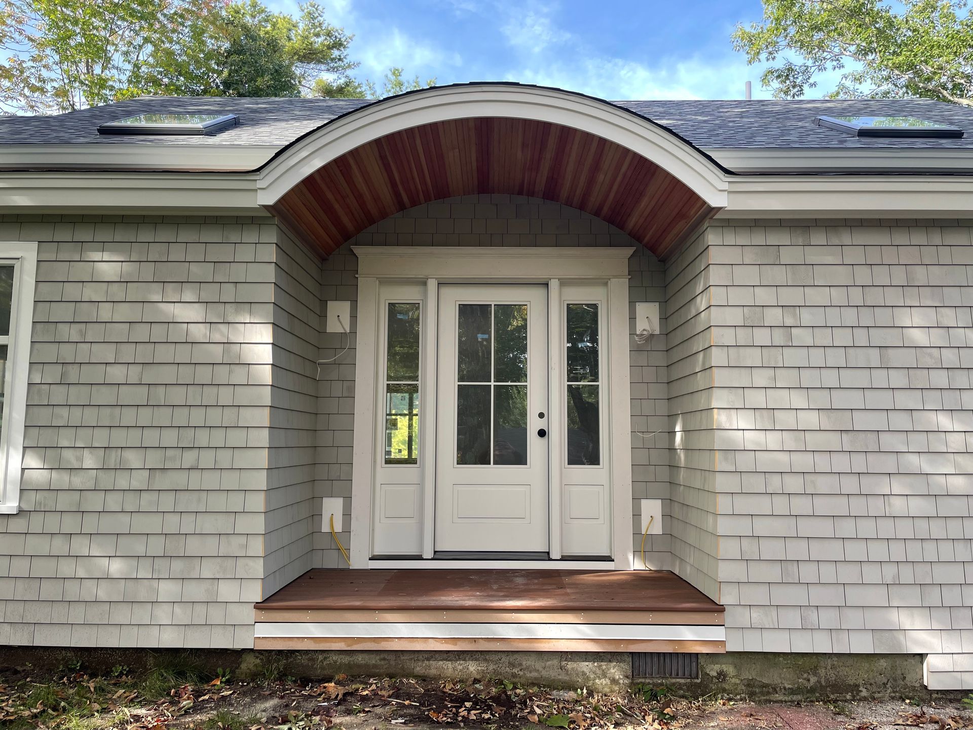 A doorway with a rounded wooden arch, centered on a gray-shingled house. Door and trim are white.