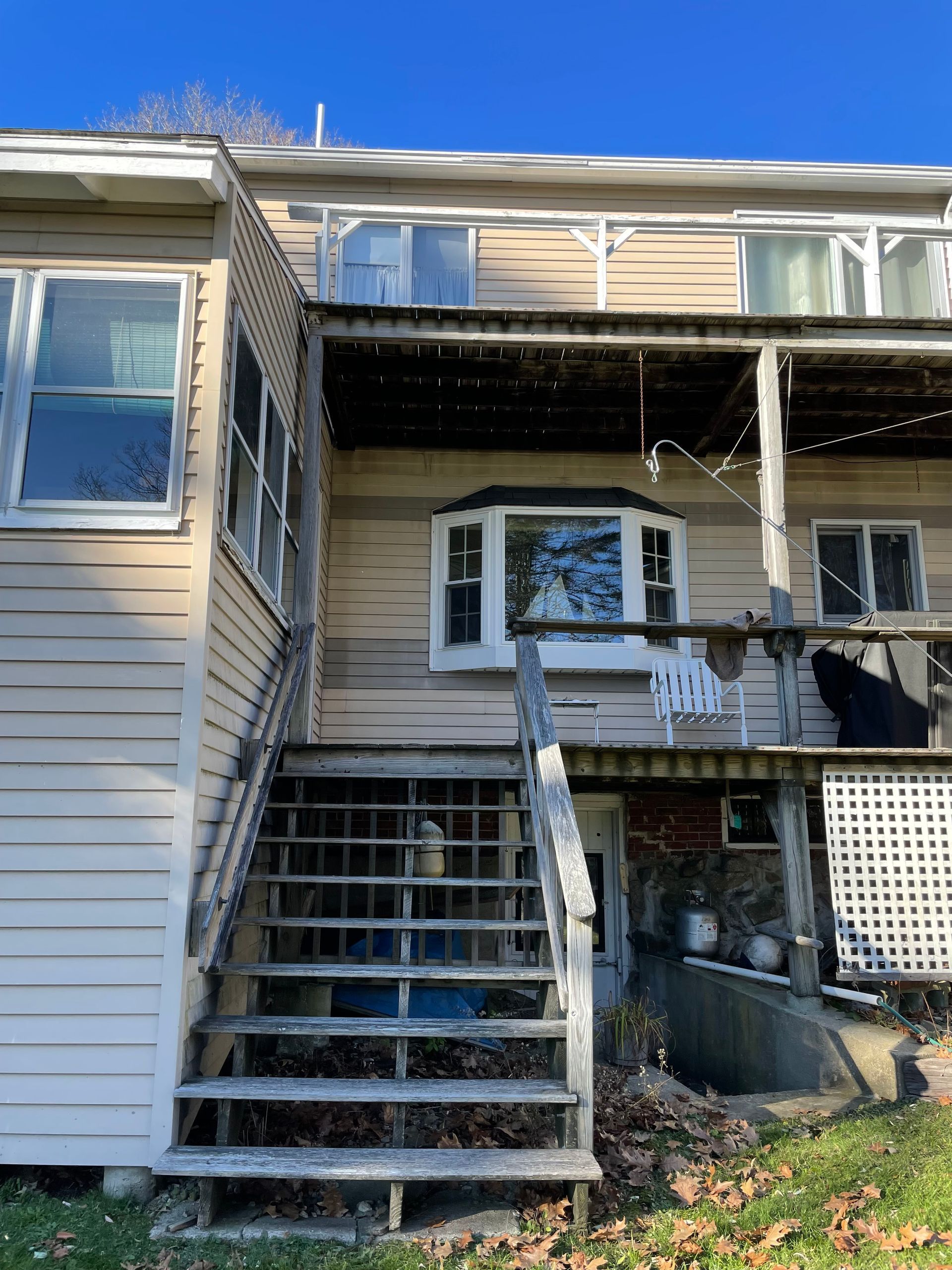 Back of a two-story beige house with weathered wooden stairs and a partially covered deck. Blue sky overhead.