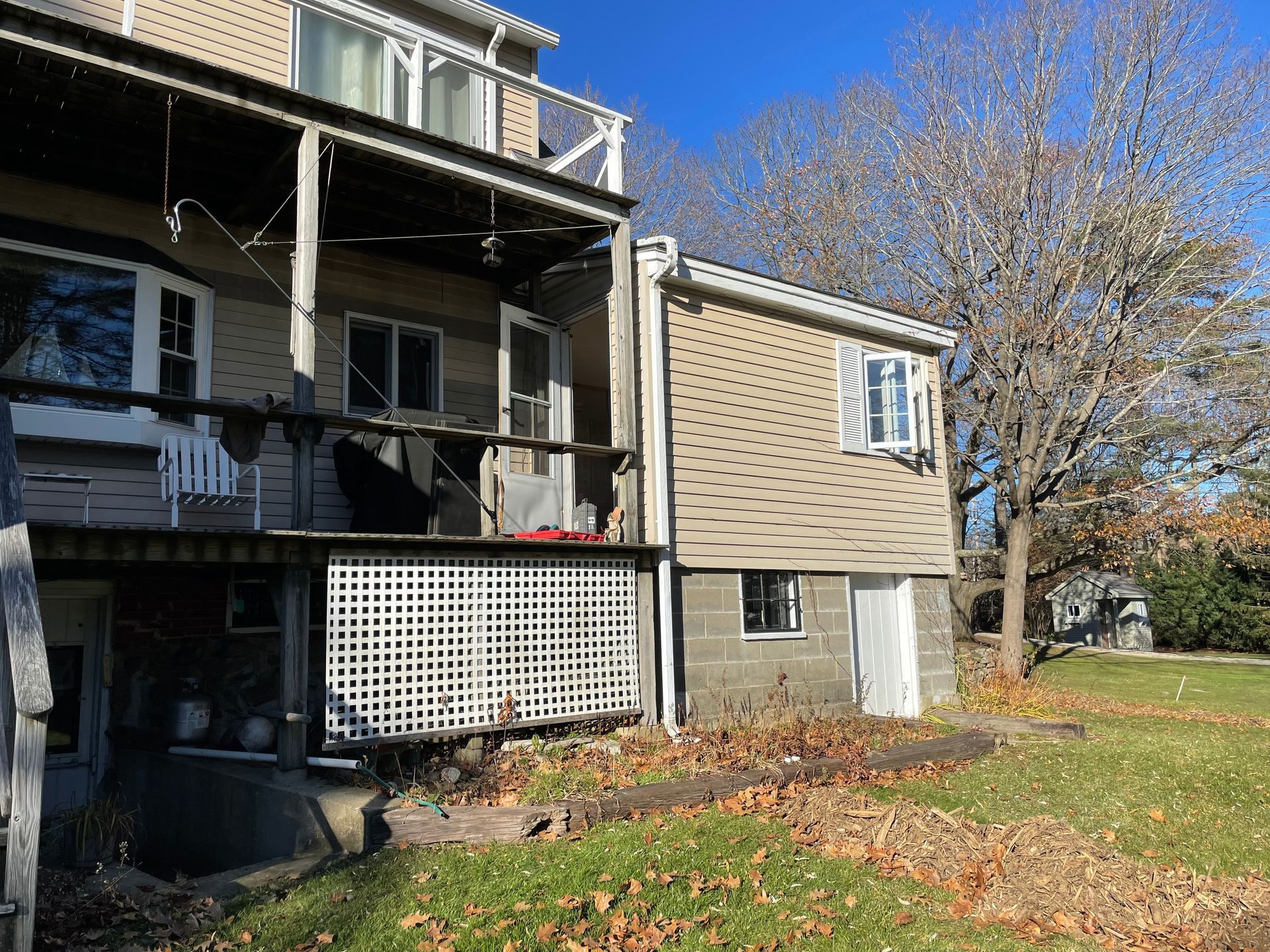 Back of a two-story beige house with a porch and a small yard with fallen leaves.