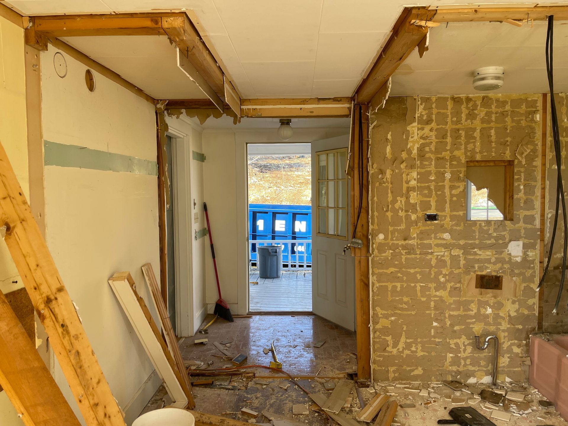 Interior demolition of a room; exposed beams, doorway, debris, torn walls, visible to outdoor area.