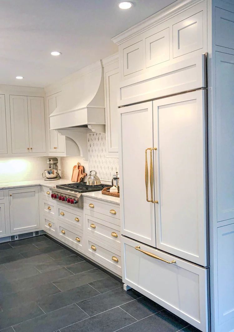 White kitchen with gold hardware, range, and built-in refrigerator. Dark gray tile floor.