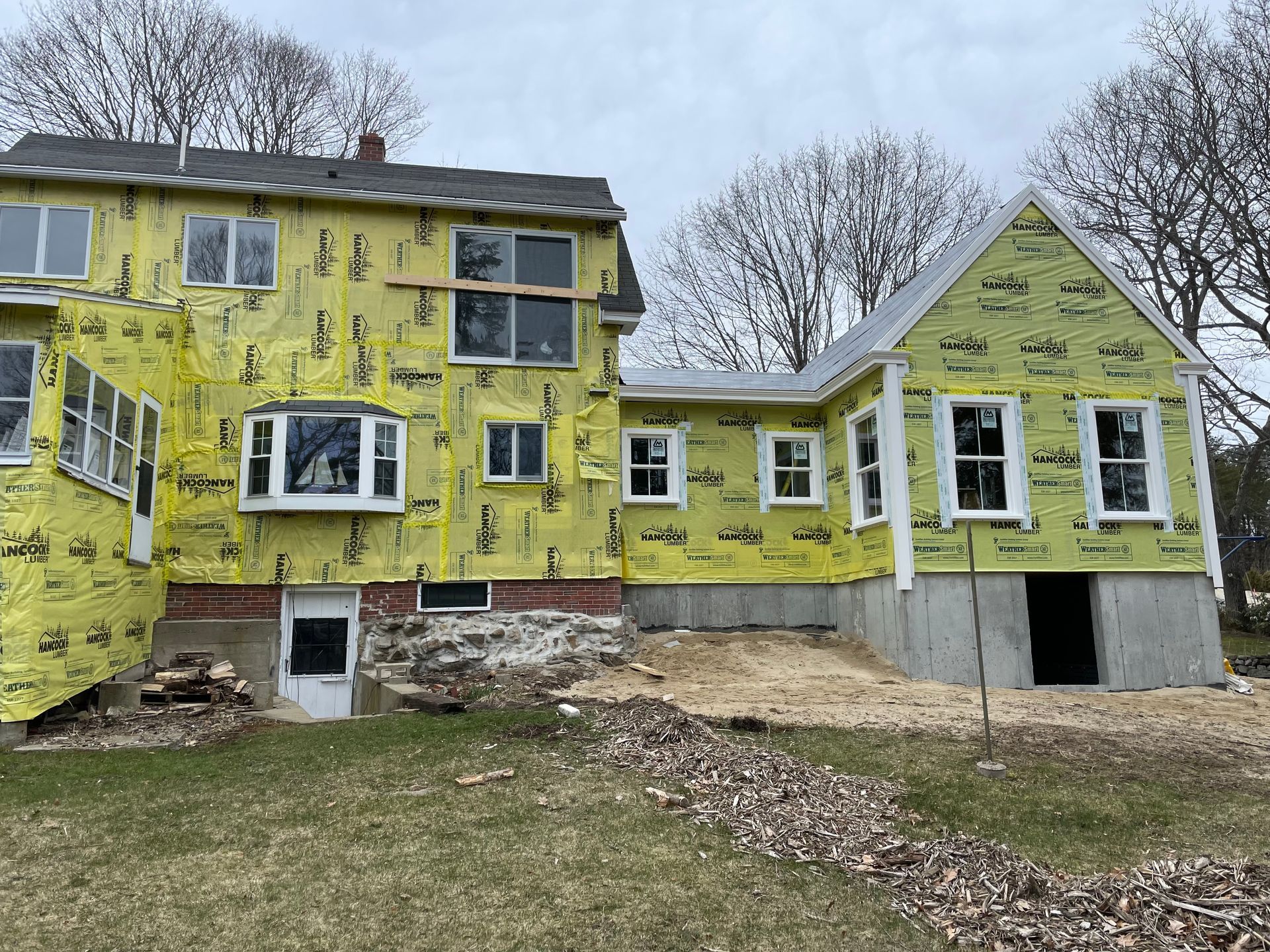 Two-story house under construction, yellow insulation, new windows installed, exposed foundation, cloudy day.