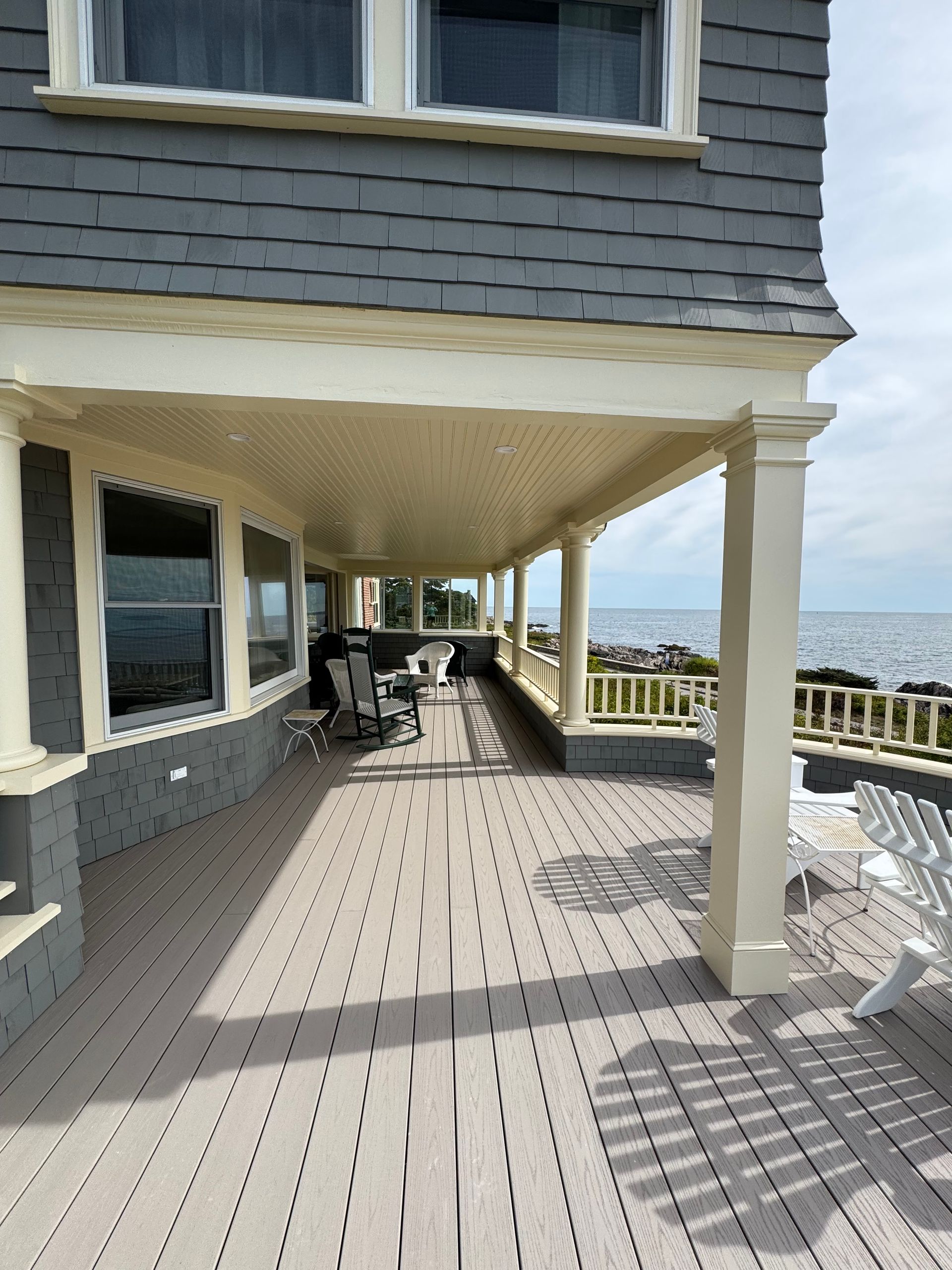 Gray house with a covered porch overlooking the ocean, white trim, wooden deck, and Adirondack chairs.