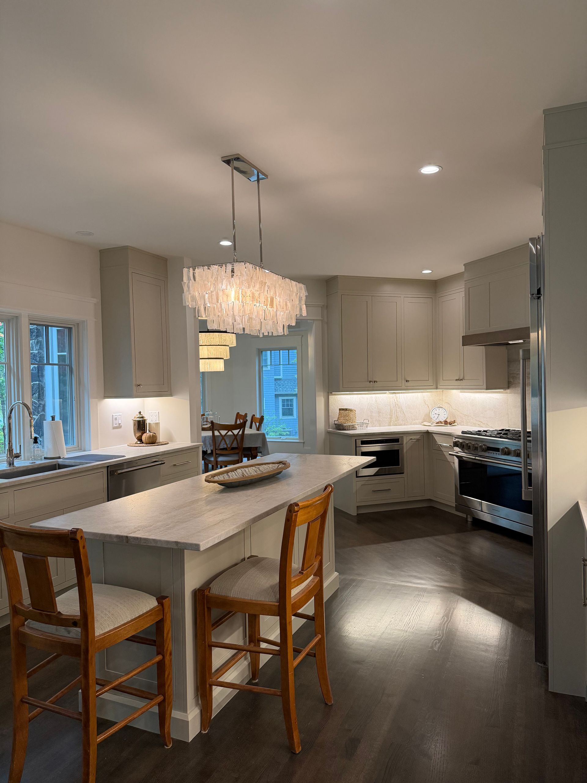 A modern kitchen with a white island, wooden chairs, and a decorative light fixture. Dark wood floors.