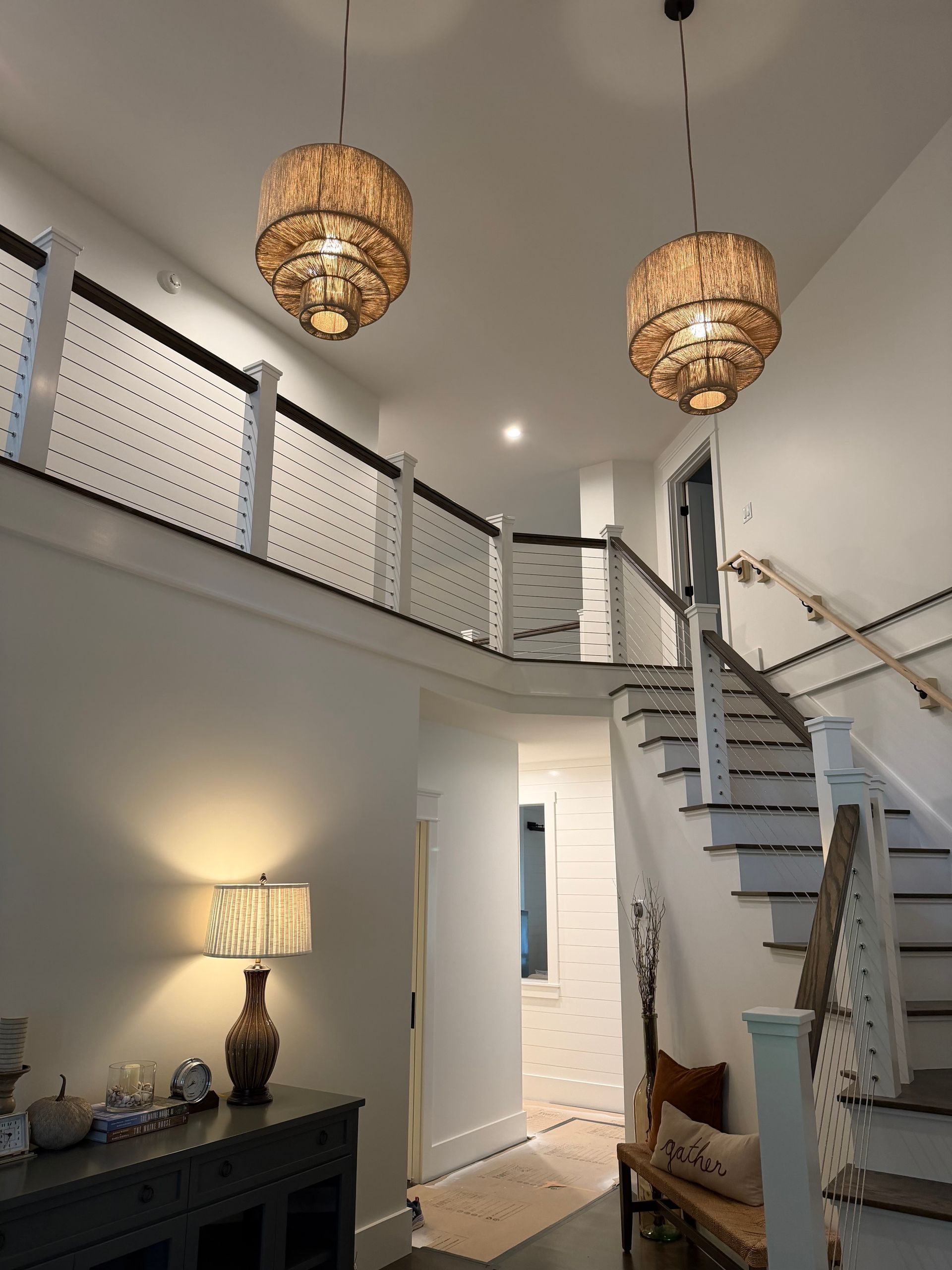Two-story entryway with chandeliers, staircase, and a table lamp. White walls, dark railings, and a wooden console.