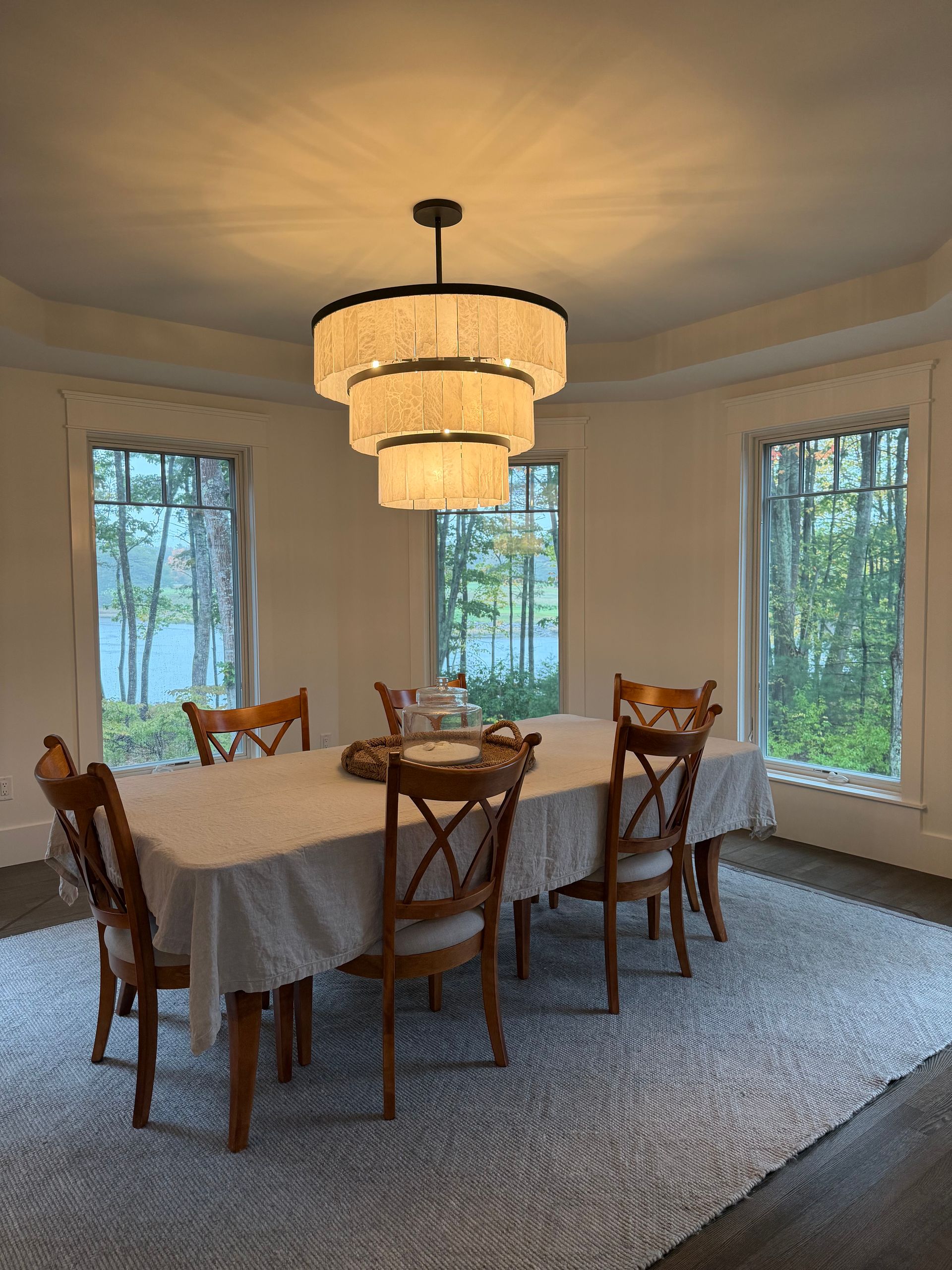 Dining room with chandelier, table set for six, surrounded by windows with outdoor view.