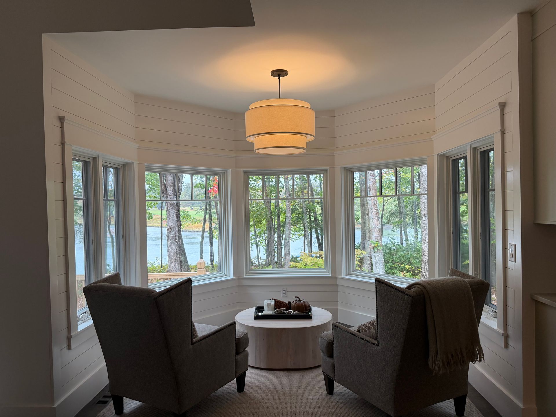 Cozy nook with two armchairs facing windows overlooking a lake. A round coffee table and a hanging light fixture complete the space.