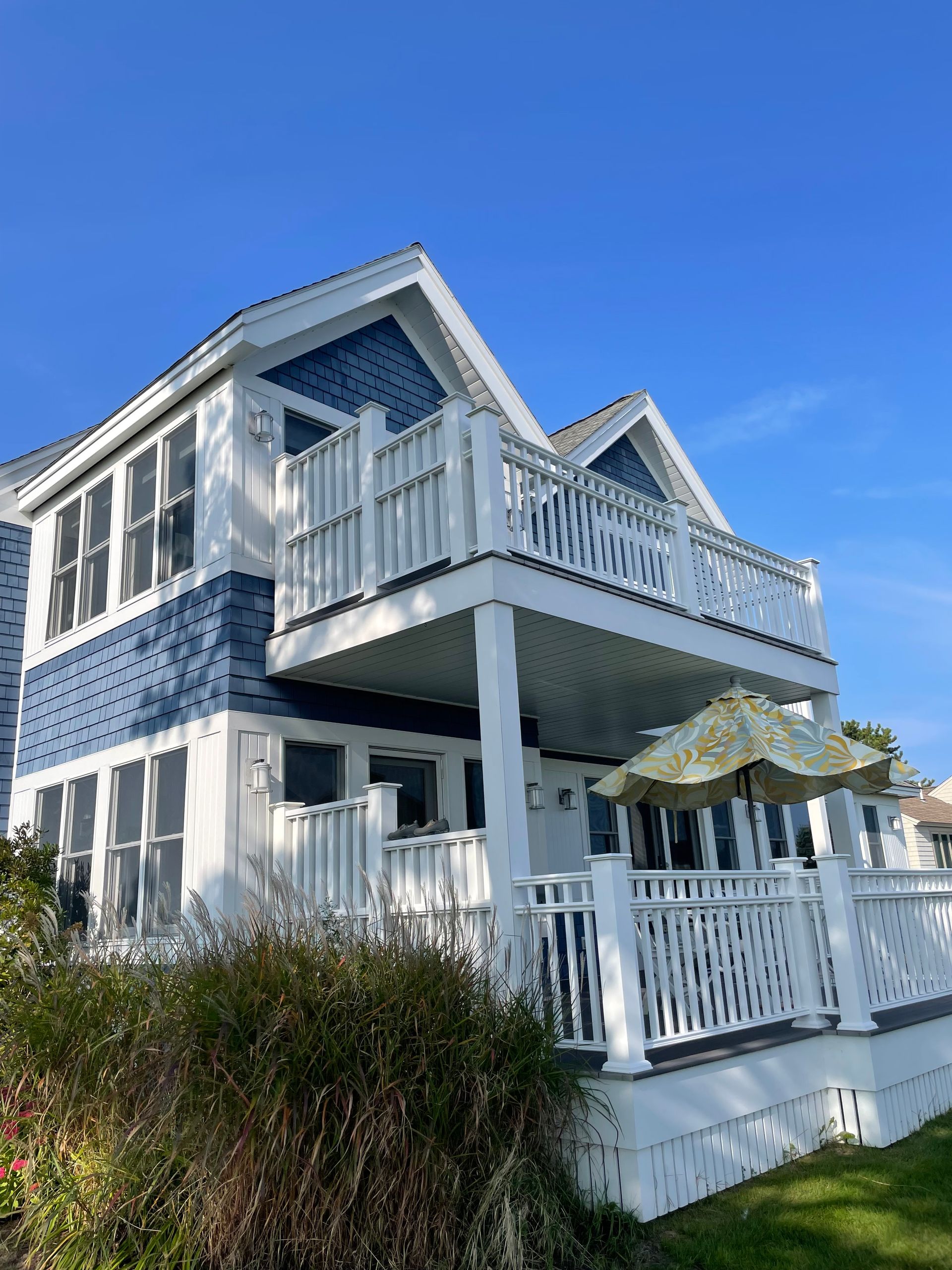 Two-story white house with blue siding, decks, and a green and yellow umbrella on the lower deck under a blue sky.