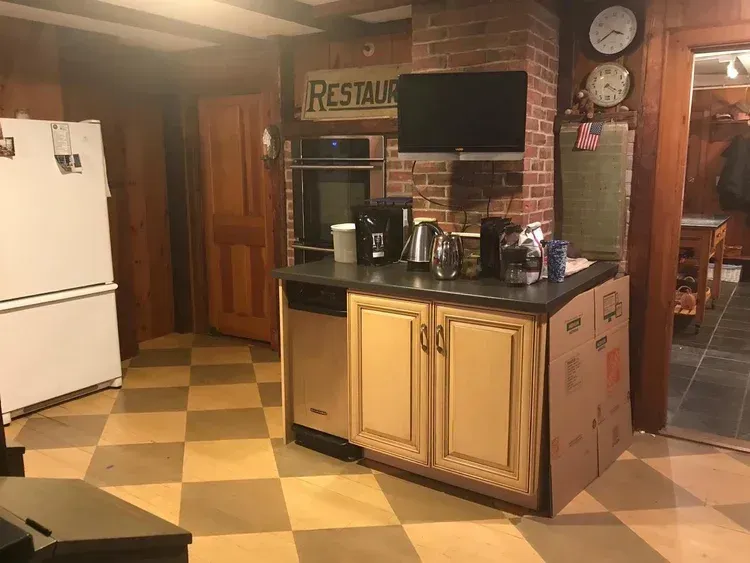 A kitchen with checkered floor. Appliances include a refrigerator, oven, dishwasher, and coffee maker. A TV hangs on brick.