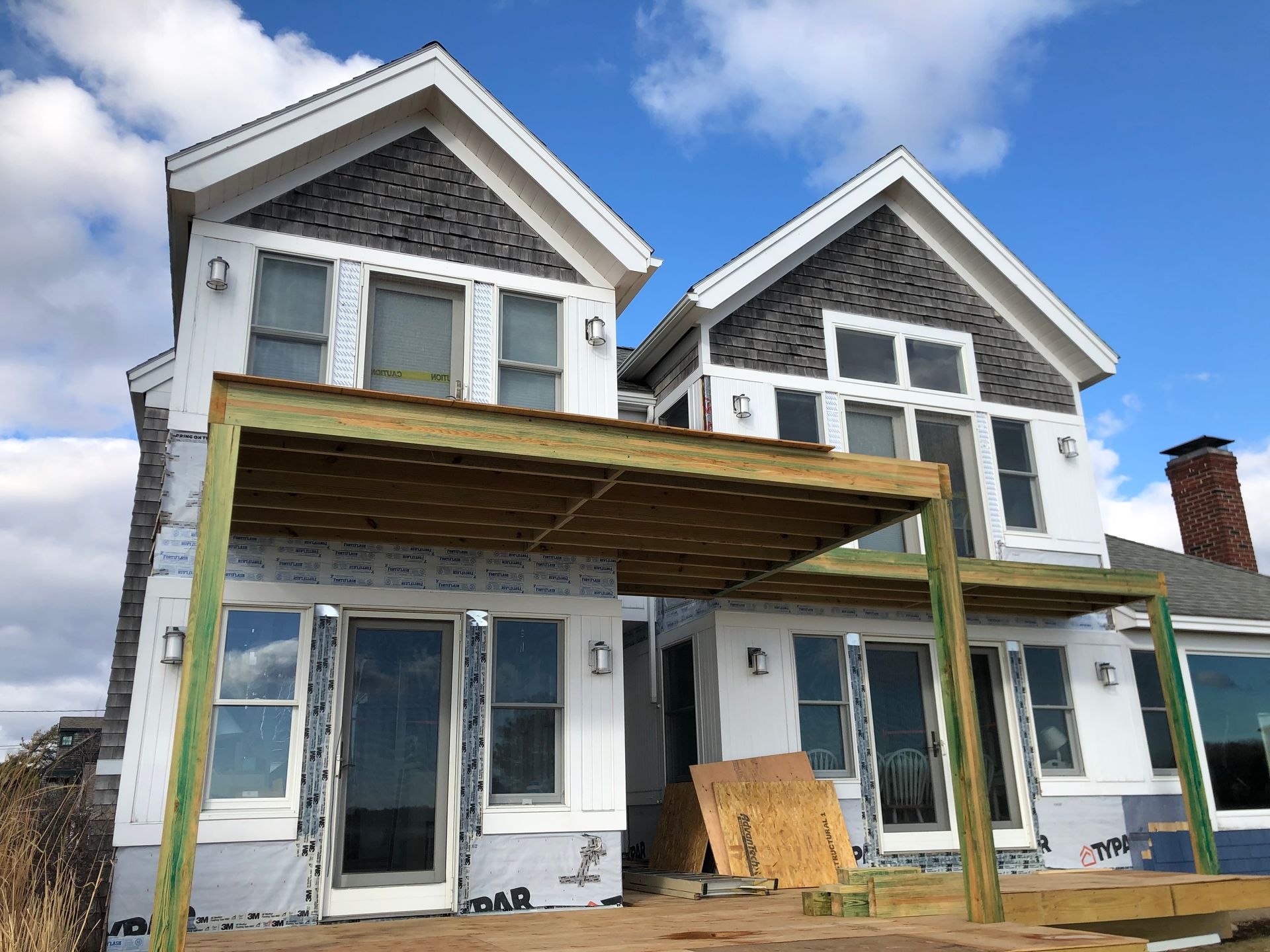 Two-story house under construction with a wooden deck; gray shingles, white siding, blue sky.