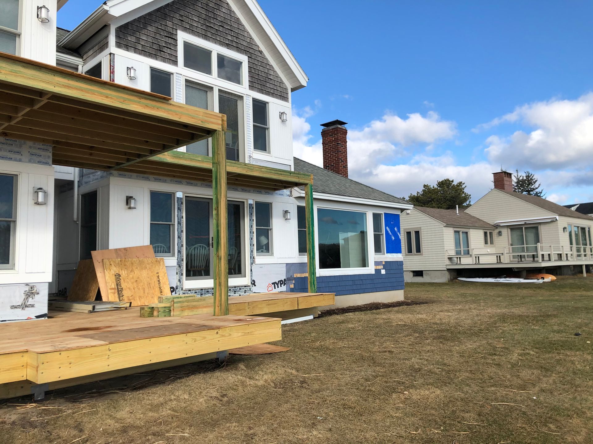 House with wooden deck under construction. Blue sky, grass, and other houses are visible.