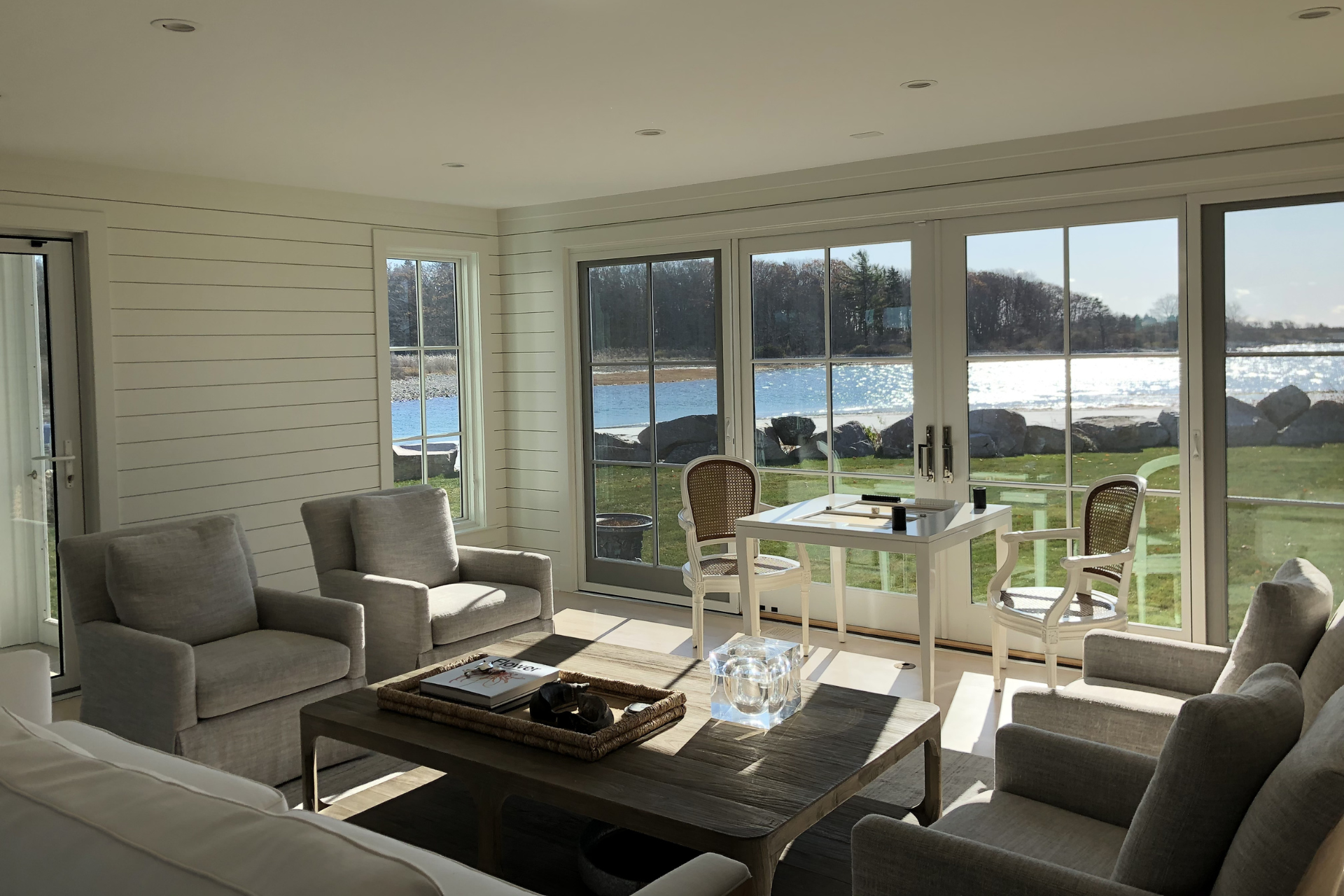 Sunlit living room with water views, gray furniture, and a small table with two chairs.