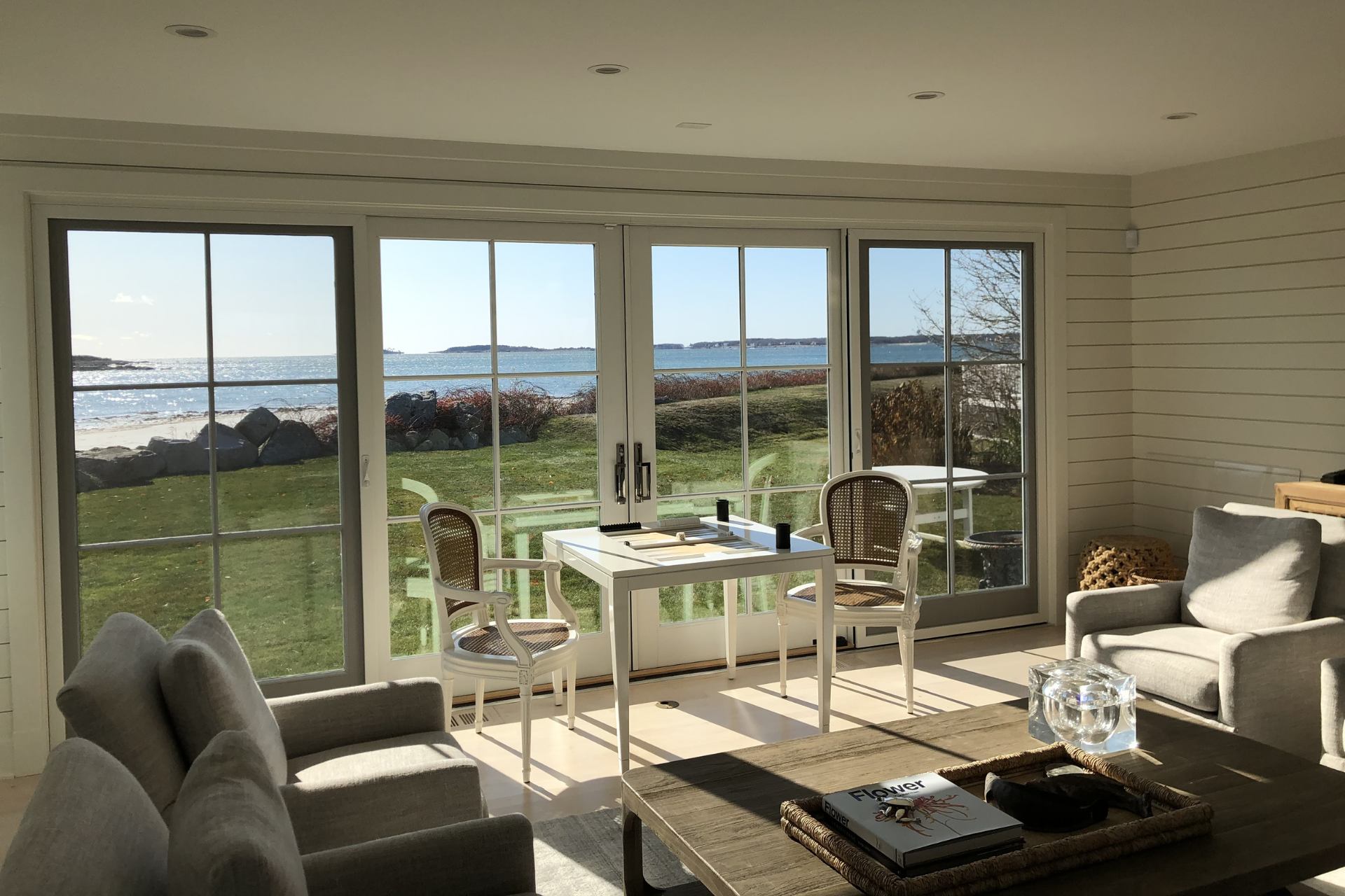 Bright interior with a view of the ocean through large glass doors; white walls, neutral furniture, and a table and chairs on the lawn.