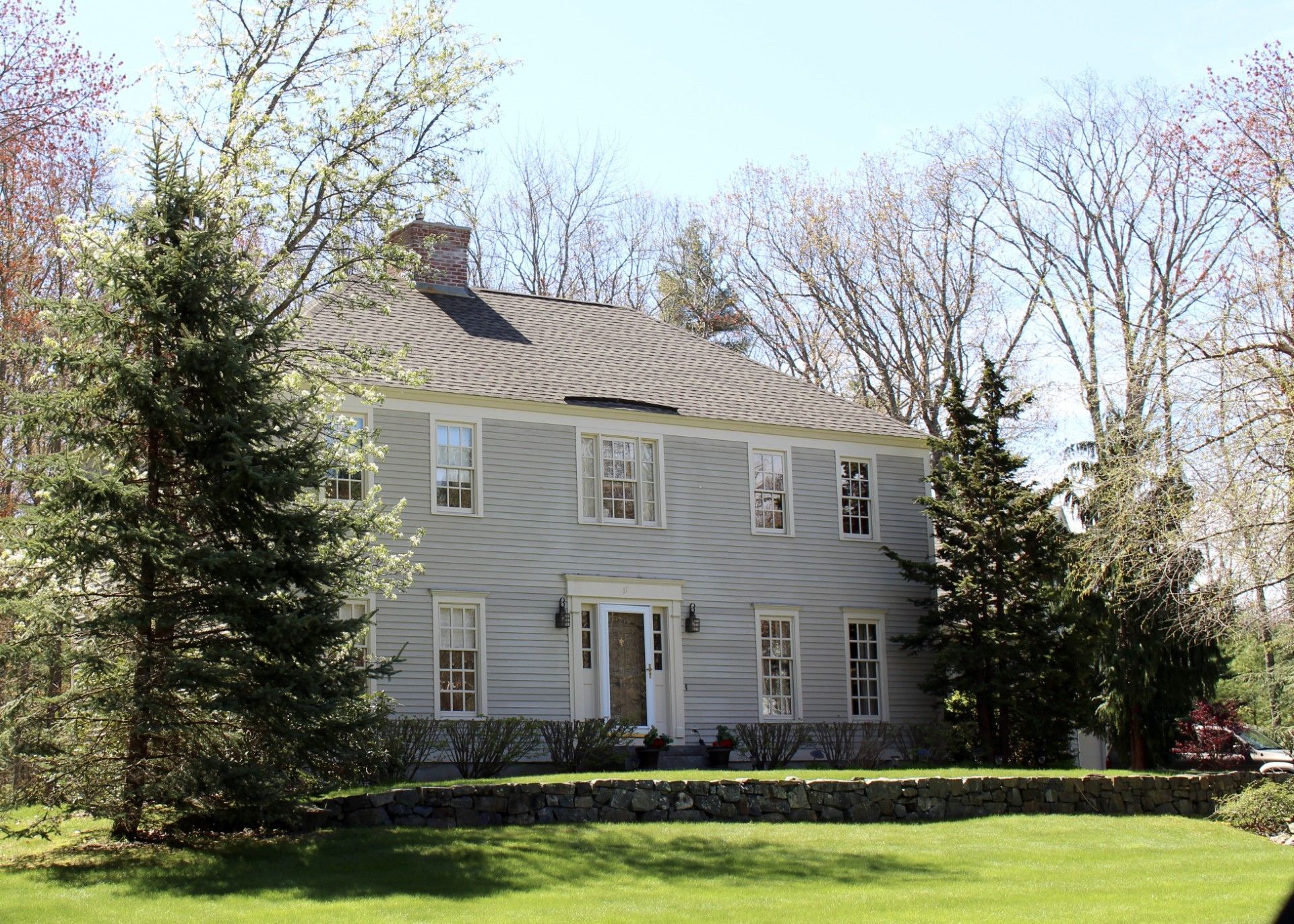 Two-story, light blue house with white trim, a stone wall in front, and trees on either side.