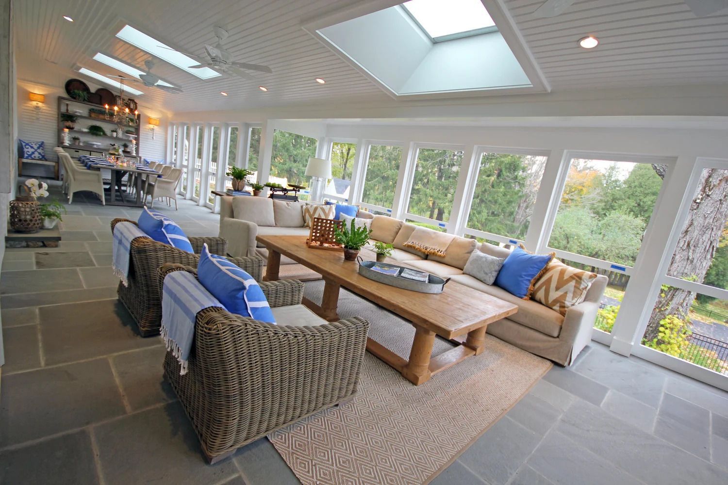 Sunroom with stone floor, wicker chairs, beige sofa, wooden coffee table, and large windows overlooking a green landscape.