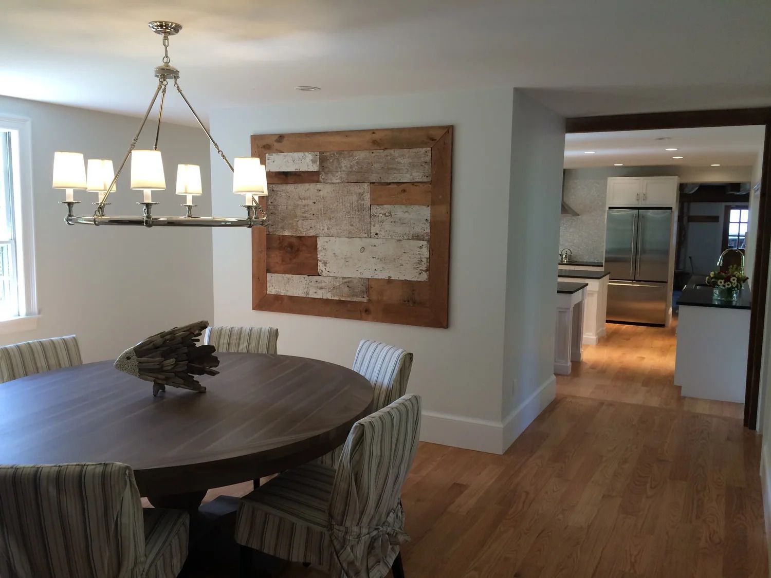 Dining room with round table, chairs, and rustic wall art. Kitchen visible in the background.