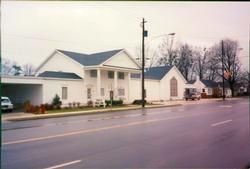 White building with blue roof beside a road, with a few cars and bare trees under an overcast sky.