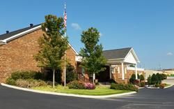Brick office building with trees and landscaped entrance beside a parking lot under a clear blue sky
