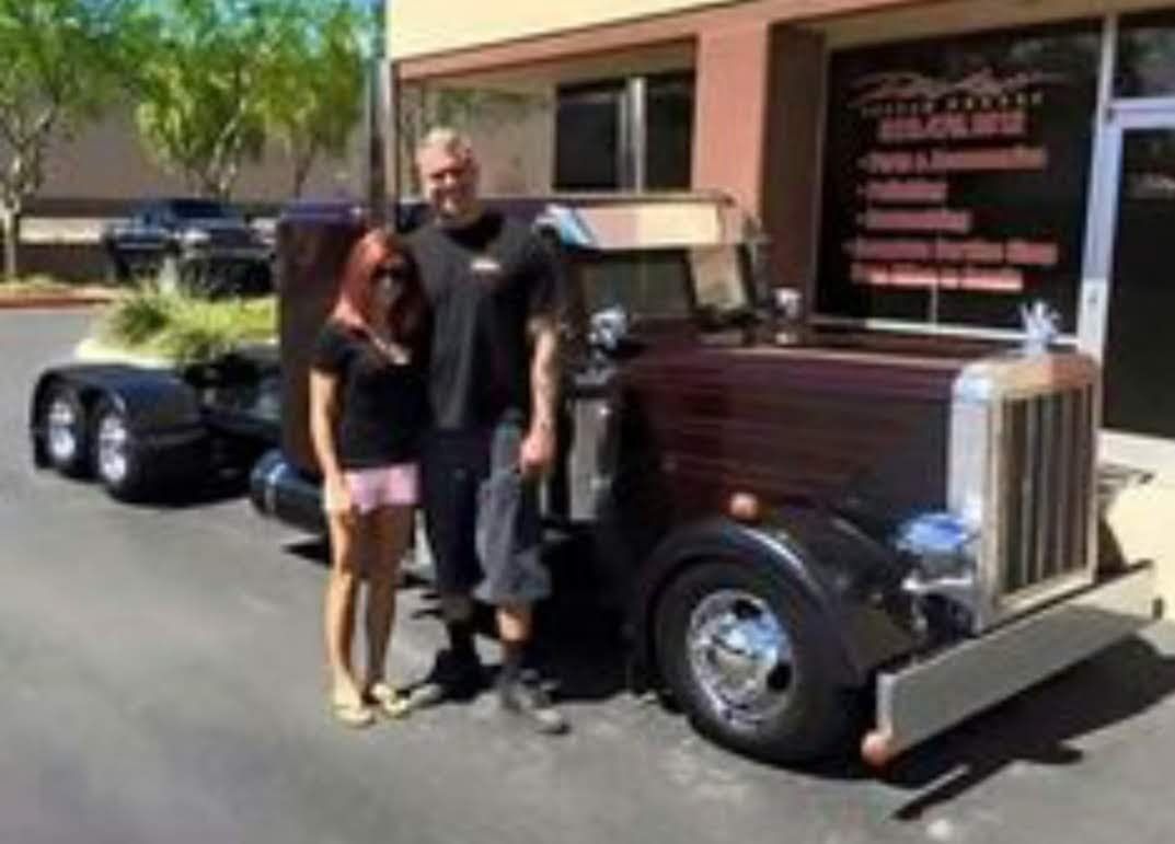 Man and woman pose with a dark red custom semi-truck in front of a business.