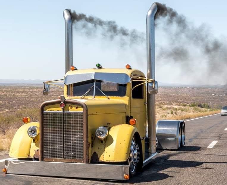 Yellow vintage semi-truck, with smoke billowing from tall stacks, driving on a desert highway.