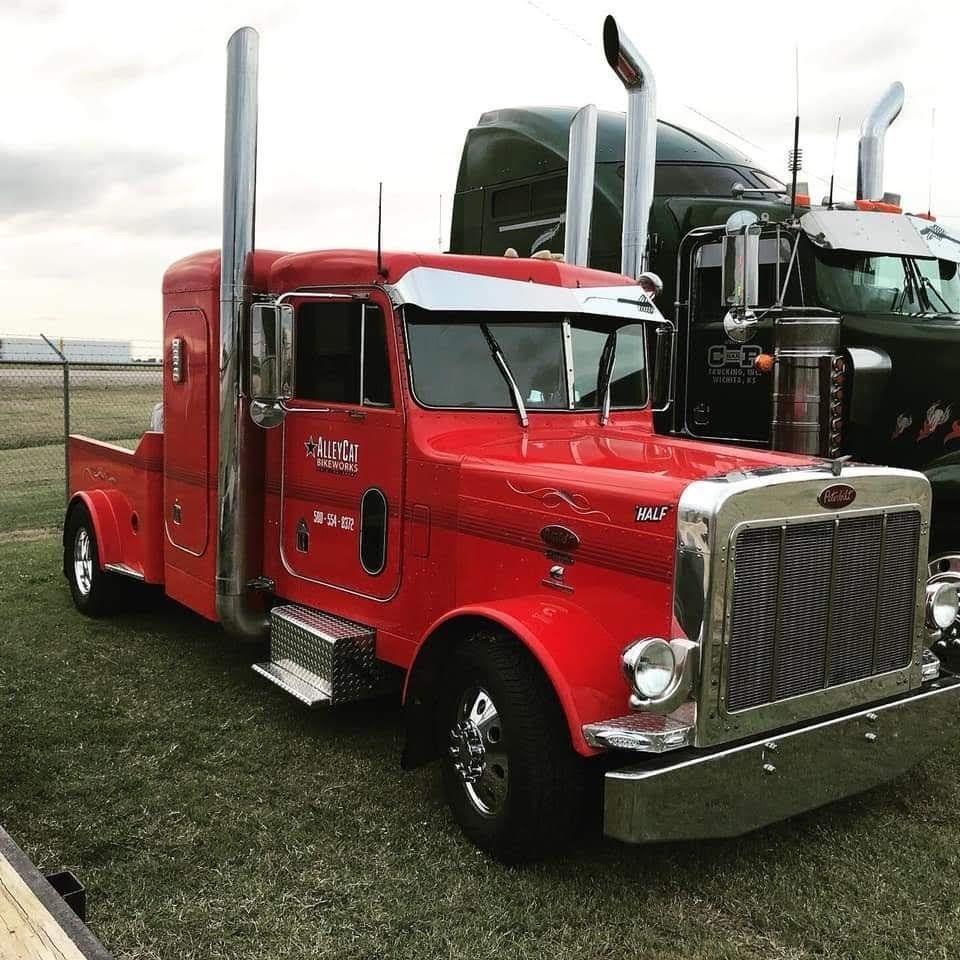 Red Peterbilt truck with chrome accents, standing on grass, another truck in background.