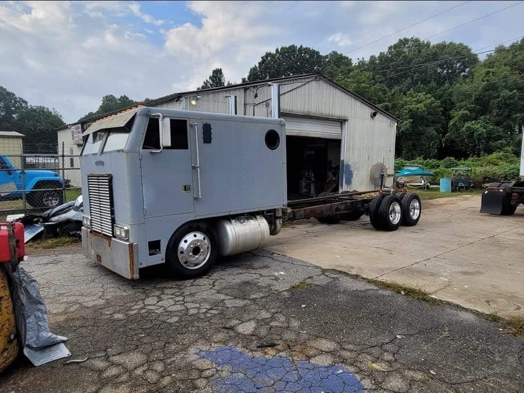 Gray semi-truck cab and chassis, missing parts, parked outside a dilapidated building.
