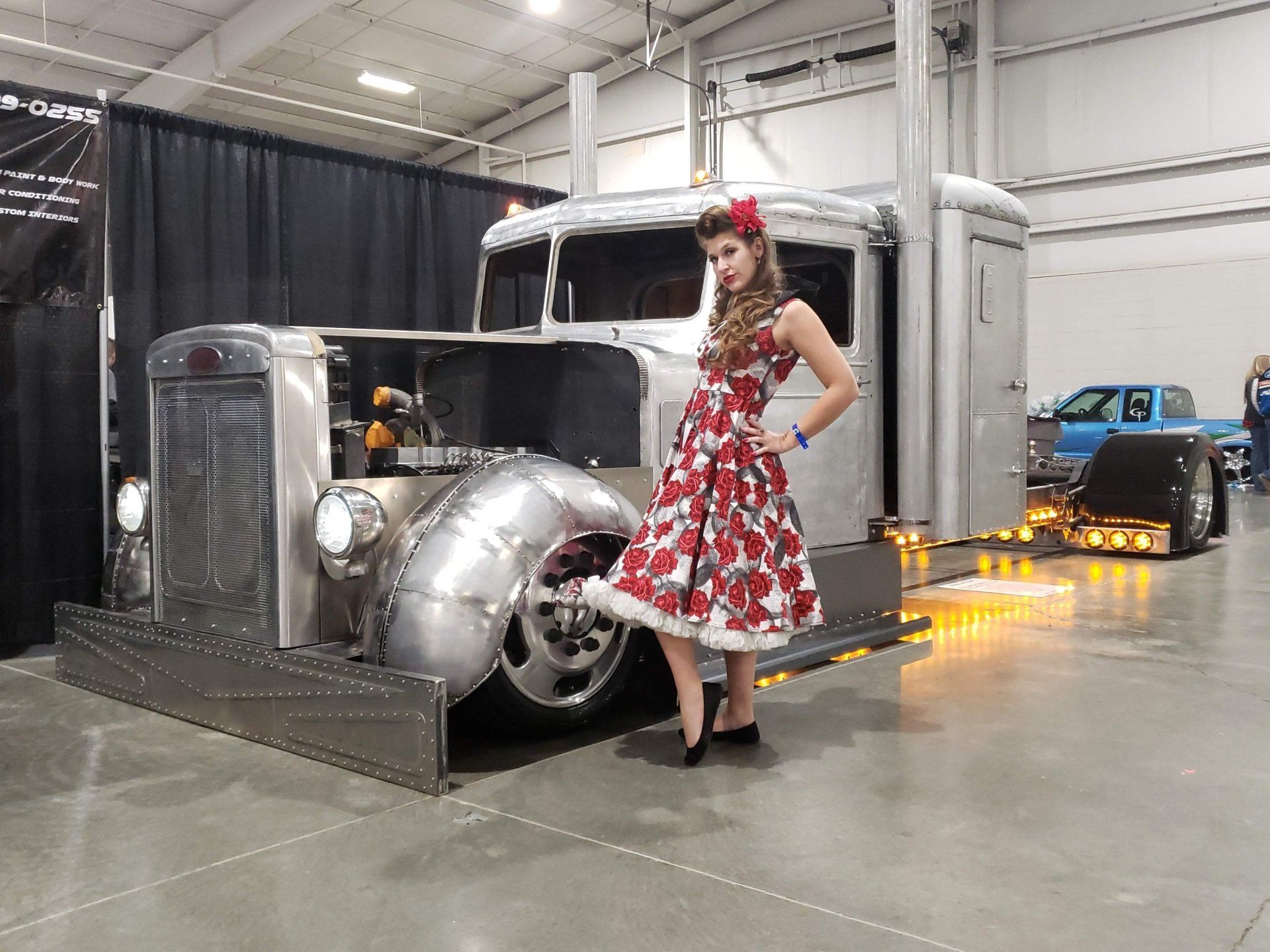Woman in floral dress poses next to a custom, silver semi-truck at an indoor car show.