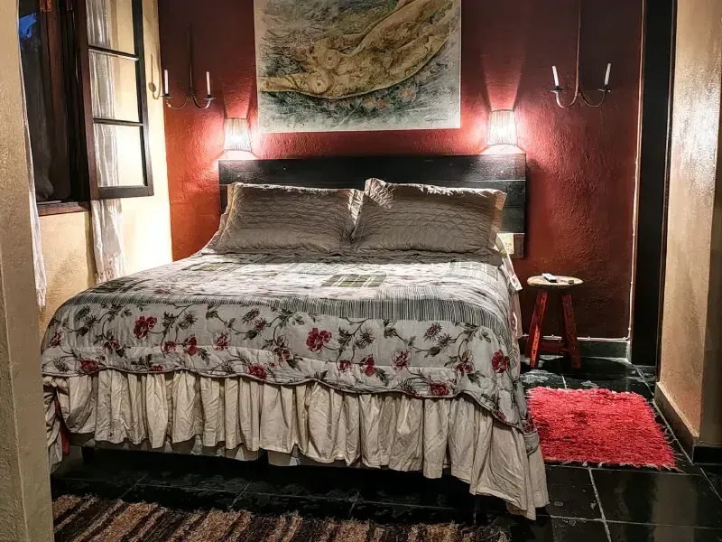 Quarto aconchegante com colcha floral, paredes de madeira escura, obras de arte na parede e um pequeno tapete vermelho.