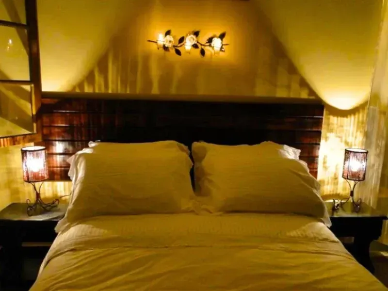 Quarto com iluminação aconchegante, cama grande, paredes amarelas, abajures e uma luminária de parede acima da cabeceira.