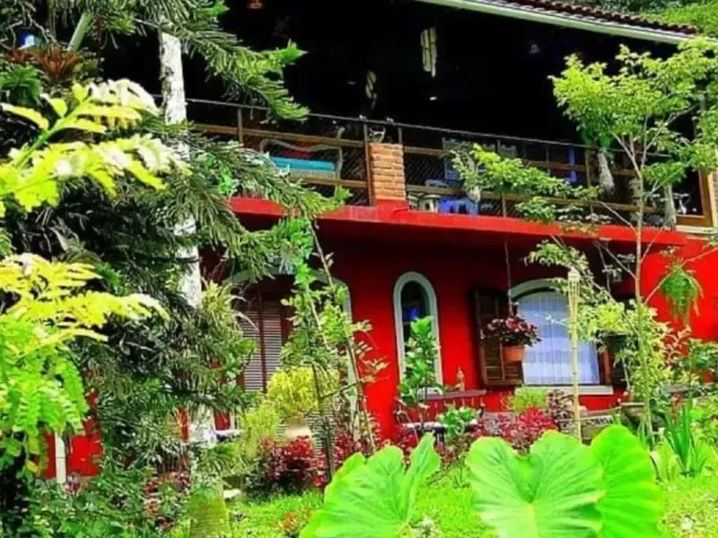 Casa vermelha vibrante com plantas tropicais verdejantes e uma varanda com vista para o jardim.